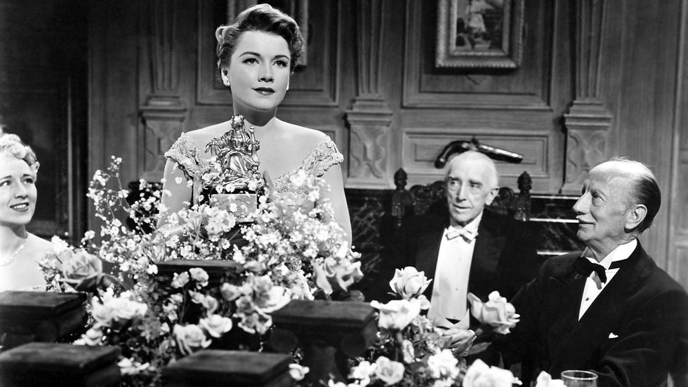 A woman holds a trophy, surrounded by flowers at a formal event. Three elegantly dressed people sit nearby in a wood-paneled room.
