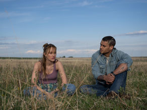 A man and woman in casual clothing sit in a field. Mood is thoughtful.