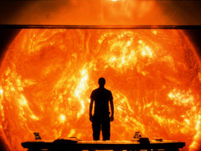 Silhouette of a person stands facing a massive, fiery sun. The room is dimly lit, creating a dramatic and intense atmosphere.