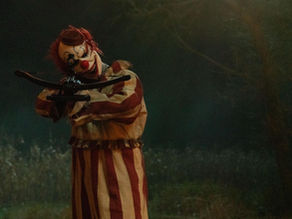 A clown in a striped costume aims a crossbow in a dimly lit, eerie forest setting at night. The scene feels tense and unsettling.
