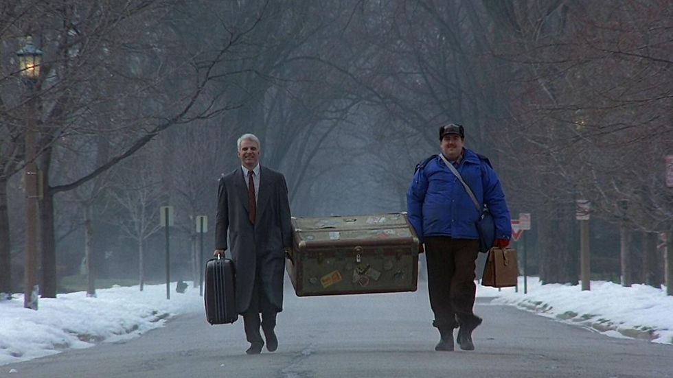 Two men walk on a snowy street, carrying a large trunk. One wears a suit, the other a blue coat, appearing content. Bare trees line the road.