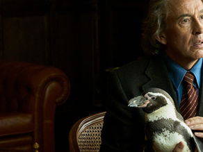 A man in a suit strokes a penguin on his knee