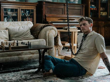 Young man in a beige shirt sits on floor, chained to furniture, near a chessboard on a wooden table. Vintage room with piano in back.