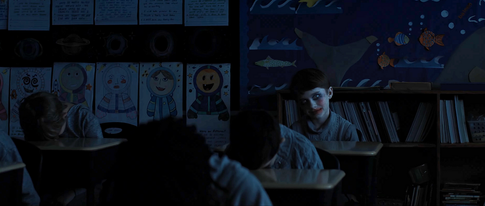 Boy with painted face in dark classroom; classmates asleep. Bright wall art of astronauts and ocean animals in background; eerie mood.