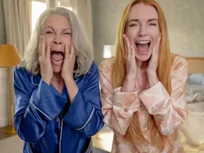Two women in pajamas scream with hands on cheeks in a bedroom. One wears blue, the other pink. Background shows a bed and lamp, conveying surprise.