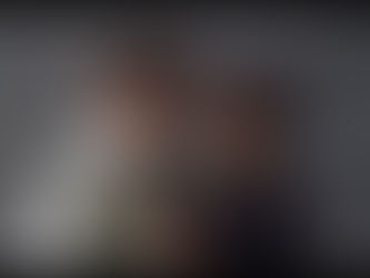 A couple stands close against a gray backdrop, gazing at each other with affection. The man in glasses wears a light shirt; the woman, a dark top.