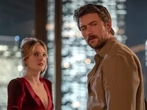 A man and a lady in a red dress with a background of skyscrapers, look off camera in a serious fashion.