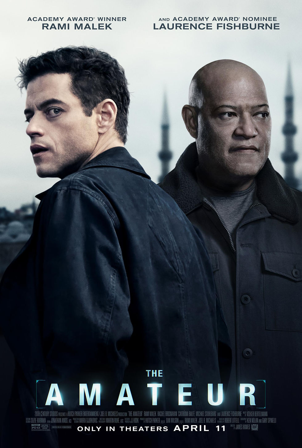 Two serious men in dark coats stand against a cityscape background. Text reads "The Amateur." Premiere date: April 11. Dark, tense mood.