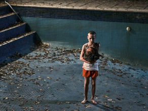 A child in an empty, leaf-filled pool holds a cat, wearing an orange outfit. The setting is dimly lit, creating a somber atmosphere.