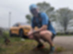 Man in blue squatting, smiling, wearing a cap and backpack. Yellow car behind him, with greenery and overcast sky in the background.