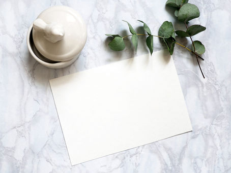 White ceramic dish with lid on marble surface, blank paper sheet beside, green eucalyptus branch above, creating a serene, minimal scene.