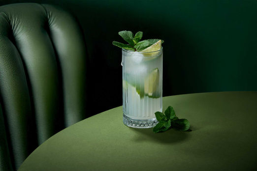 A crystal glass of mojito with ice, lime wedges, and mint leaves on a green table, photographed by a professional drinks photographer  in sussex with elegant lighting and styling.
