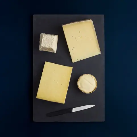Overhead shot of a selection of cheeses and a cheese knife arranged on a black slate board, set against a dark blue background. The cheeses vary in shape and texture, from hard blocks to a soft, round cheese in a wooden container. Food and drink photographer Brighton, Sussex, Brighton food photographer, Stuart Price.