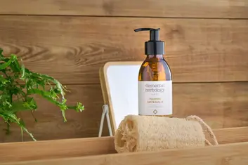 A bottle of Elemental Herbology Rejuvenate Bath & Body Oil is elegantly displayed on a rolled natural loofah, set against a wooden spa-like backdrop. A small mirror with a wooden frame and a green plant add to the tranquil, organic aesthetic. Captured by product photographer, Stuart Price in Brighton, Sussex, this wellness-themed composition highlights luxury skincare in a warm, natural setting.