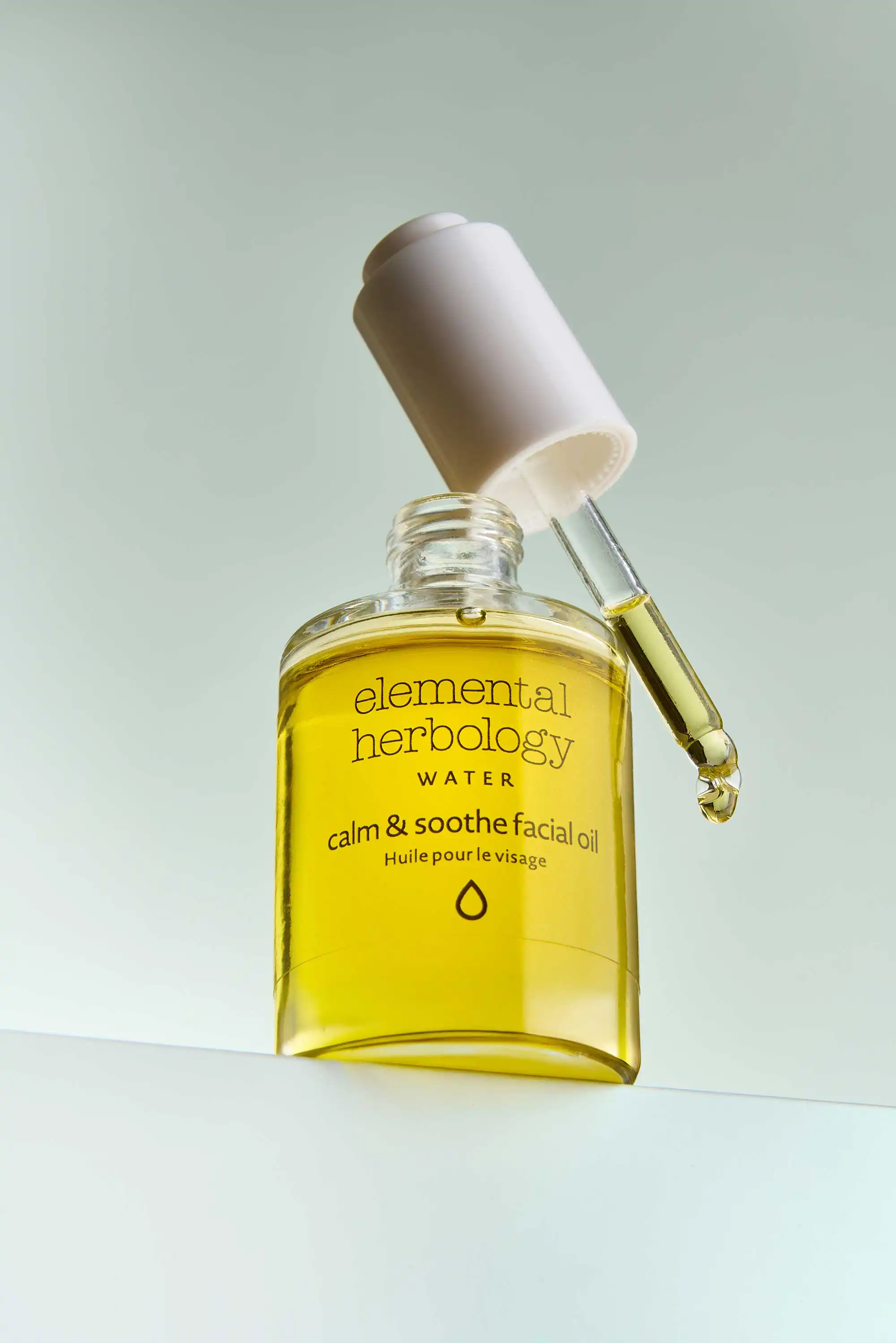 A product photograph by a Sussex product Photographer features a clear, square glass bottle of Elemental Herbology Calm & Soothe Facial Oil. The golden-yellow oil is visible inside the bottle. The white dropper cap is slightly ajar, with a drop of the oil clinging to the end of the glass pipette. The label is white with black text, clearly stating "elemental herbology" at the top, followed by "WATER," "calm & soothe facial oil," and "Huile pour le visage." A minimalist circular symbol is at the bottom of the label. The bottle sits on a reflective surface against a soft, muted green background. The lighting is bright and even, highlighting the clarity of the oil and the simple elegance of the packaging.
