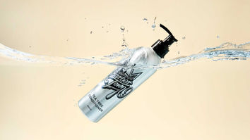 The image shows a dynamic product photograph featuring a silver Barber Town Tea Tree Conditioner bottle partially submerged in water with a splash effect. The bottle is positioned horizontally with water flowing around it against a neutral beige background. The black pump dispenser is visible, and water droplets are suspended in the air, creating a sense of movement and freshness that emphasizes the product's hydrating qualities. This creative commercial photography demonstrates advanced liquid splash photography techniques typical of high-end product photography. The composition would be suitable for advertising, e-commerce, or social media marketing by Stuart Price, a professional product photographer based in Brighton or Sussex who specializes in creating eye-catching product imagery with technical precision and creative flair.