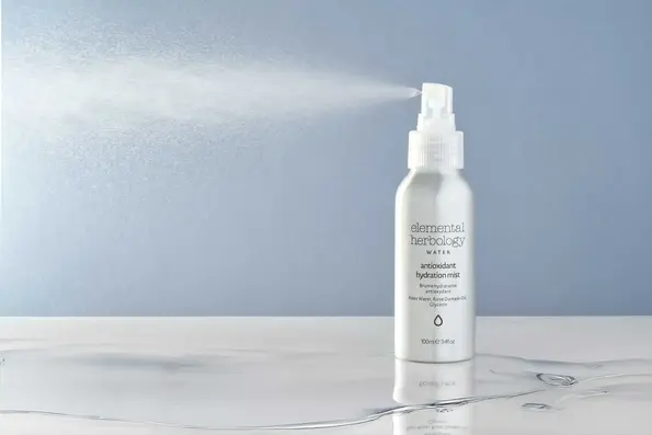An aluminium can of Elemental Herbology Antioxidant hydration mist spraying mist in the air against a light blue background.  Captured by product photographer, Stuart Price in Brighton, Sussex, the mist is lit from behind showing a fine spray. The can is on a marble surface covered in a small puddle of water.