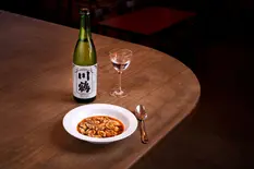 This image showcases a carefully composed Japanese dining scene featuring a traditional pairing of sake and seafood. The photograph presents:

A white ceramic bowl containing what appears to be a seafood stew or soup with small shellfish (likely clams or mussels) in a rich, reddish-orange broth, topped with fresh herbs. The deep bowl allows the vibrant colors of the dish to stand out against its clean white interior.

A bottle of premium sake with a distinctive black and white label featuring large Japanese calligraphy characters (川鶴 - Kawatsuru), which indicates this is likely from the Kawatsuru brewery, one of Japan's oldest sake producers (the label notes "SINCE 1891").

A stemmed glass partially filled with clear sake, positioned between the bottle and the food, creating a visual triangle that draws the viewer's eye across the composition.

A silver spoon placed beside the bowl, adding a touch of elegance and practicality to the setting.

The entire arrangement sits on a warm-toned wooden round table with visible grain, which provides a natural, organic backdrop that complements the food and drink. The background is deliberately darkened, creating a moody, intimate restaurant atmosphere with hints of other tables visible.

The lighting in this image is particularly notable - a focused, dramatic spotlight effect that illuminates the food and drink while allowing the surroundings to recede into shadow. This lighting technique is characteristic of Stuart Price's food photography style from Brighton, Sussex, emphasizing the textures and colors of the cuisine while creating an intimate dining atmosphere.