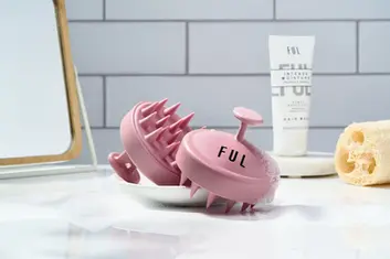 A product photograph by a Brighton product photographer showcases two pink silicone scalp massager brushes from FUL on a white, slightly textured surface. One brush is upright, displaying the black "FUL" logo on its handle and its soft silicone bristles. The other brush rests on its side, revealing more of its bristle detail. Behind the brushes, a white tube of FUL Intense Moisture Hair Mask stands upright. To the right, a natural-colored loofah sponge adds a textural element. The background features light grey, horizontally stacked tiles and a blurred reflection of a wooden-framed mirror on the left. The lighting is soft and bright, highlighting the textures of the different products in this bathroom setting.