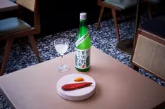 This image captures a minimalist Japanese dining scene with a focus on simplicity and fine presentation. The composition features:

A distinctive bright green sake bottle with traditional Japanese labeling and calligraphy, featuring folk art-style illustrations and a white cap. The bottle appears to be a specialty nigori (cloudy) sake or possibly a seasonal release.

A stemmed wine glass partially filled with clear sake, positioned to the left of the bottle.

A modern, circular white serving plate containing two elements: a piece of red-coated fish (likely tuna or salmon with a spice coating) and a small orange garnish or pickled vegetable.

The setting includes a light pink or neutral table surface contrasted against a black and white terrazzo or speckled floor pattern. Mid-century modern furnishings are visible in the background, including wooden chair legs and what appears to be a vintage speaker or audio equipment with cane detailing.

The lighting creates a moody atmosphere with dramatic shadows that highlight the food and drink while maintaining the authentic ambiance of the space.

This image exemplifies Stuart Price's food and drink photography style from Brighton, Sussex, with its emphasis on clean composition, thoughtful lighting that enhances textures and colors, and the artful capture of Japanese culinary aesthetics within a contemporary setting. The photograph would be ideal for restaurant marketing, beverage features, or editorial content highlighting Japanese cuisine.