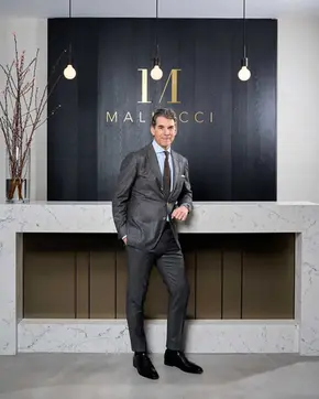 Professional corporate portrait of Patrick Mallucci in a tailored gray suit standing beside a marble counter in his Mayfair clinic interior with warm lighting. Captured by Brighton portrait photographer specializing in executive photography across Sussex.
