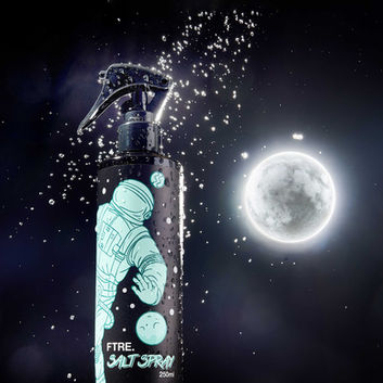 Creative studio shot of FTRE Salt Spray 250ml bottle, photographed by Brighton product photographer Stuart Price. The black spray bottle features a striking turquoise astronaut illustration with planetary graphics, positioned against a dramatic cosmic-themed background with a glowing moon and water droplets frozen mid-air. The styling and lighting emphasize the product's edgy, futuristic branding and textural surface. This high-impact product image showcases the professional work of a Sussex-based commercial photographer, ideal for marketing campaigns in the grooming and lifestyle sector.