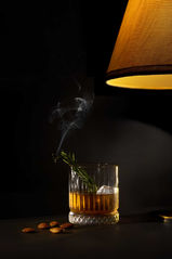 A dark, moody, low-key photograph of a whiskey cocktail in a heavy glass, garnished with a sprig of smoking rosemary. A warm, glowing lamp is visible in the upper right.