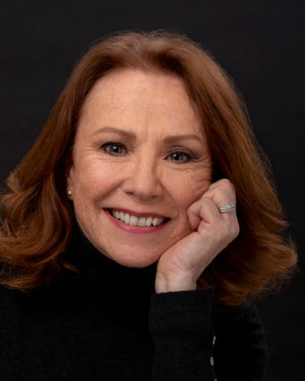 A professional portrait photograph, captured by portrait photographer in Sussex - Brighton, Stuart Price, features Melanie Hill. The close-up shot focuses on Melanie's face and upper body against a dark grey, almost black background, which emphasizes her features. Her warm, light brown hair is styled with soft waves framing her face. She has a gentle smile and her gaze is directed towards the viewer, creating a connection. Her right hand is gently resting under her chin, a classic portrait pose that adds a touch of thoughtfulness. She is wearing a dark turtleneck sweater. The lighting is soft and even, highlighting her complexion and the texture of her hair. This intimate portrait captures Melanie Hill's approachable and engaging demeanor, showcasing the skill of a Sussex portrait photographer in creating a compelling image.