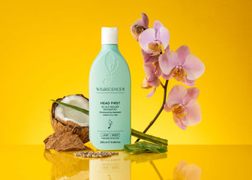 Product photography by Stuart Price, Brighton product photographer in Sussex, showcasing Wild Science Lab's "Head First Scalp Relief Shampoo." The shampoo bottle is surrounded by natural elements like coconut, orchids, aloe vera, and seeds on a reflective yellow surface.