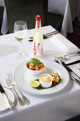 Fine dining table setting with white linens showing a plated Asian fusion dish with fried items and garnishes, Japanese sake bottle with red cap, filled wine glass, and elegant silverware arrangement at Boisdale Restaurant
