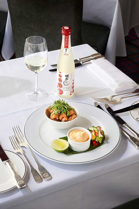 Fine dining table setting with white linens showing a plated Asian fusion dish with fried items and garnishes, Japanese sake bottle with red cap, filled wine glass, and elegant silverware arrangement at Boisdale Restaurant