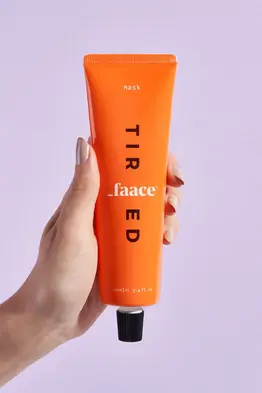 A close-up product shot of an orange tube of Faace Tired Mask, held by a hand with neatly manicured nails against a pastel purple background. The tube features bold black typography spelling "TIRED" vertically, with the brand name "_faace" in white lettering. The black screw cap is visible at the bottom. The lighting is soft and even, highlighting the sleek packaging. Captured in a clean, minimalist style, ideal for skincare branding and e-commerce. Shot by professional product photographer in Brighton, Sussex. Brighton product photographer specializing in beauty and skincare photography.