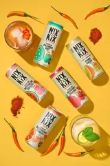 Flat-lay product photography of Nix & Kix sparkling drinks, featuring multiple colorful cans on a bold yellow background. Surrounding the cans are fresh ingredients, including red chili peppers, powdered spices, citrus slices, and mint leaves, emphasizing the brand’s signature cayenne zing. Two cocktail glasses with ice and garnishes enhance the refreshing appeal. This vibrant composition, expertly captured by Brighton drinks photographer Stuart Price, showcases premium Sussex drinks photography, perfect for beverage branding and advertising.