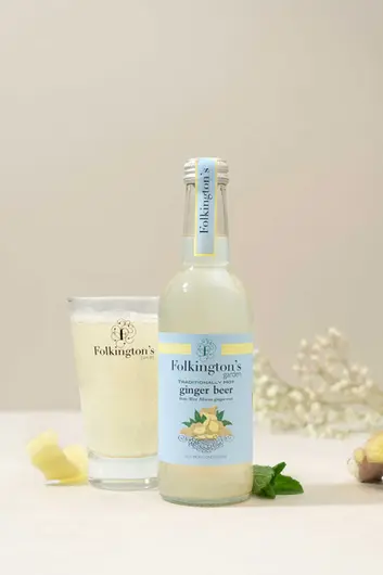The image shows a product photography setup featuring Sussex Drinks Manufacturer, Folkington's Garden traditionally hot ginger beer. The bottle is displayed with a glass partially filled with the pale yellow beverage, showing some carbonation. The pale blue label indicates it's made from West African ginger root and notes it's "not from concentrate." The composition includes decorative elements like ginger pieces, mint leaves, and small white flowers in the background, all arranged on a clean, neutral surface. This professional drinks product photography would be suitable for marketing materials, e-commerce listings, or social media.