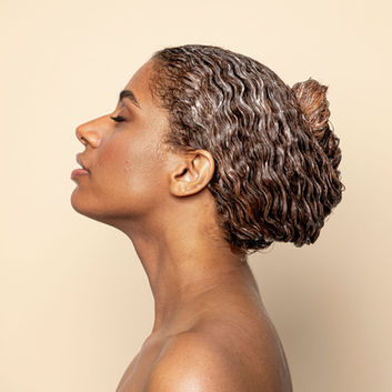 Side profile beauty portrait of a model with wet, curly hair styled in a bun against a beige background, showcasing natural skin texture and elegant profile. Professional photography by Sussex and Brighton model photographer.