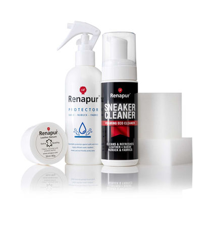 A product photograph by a Sussex product photographer showcases a set of Renapur cleaning and protection products. From left to right, there is a white tub of Renapur Leather Balsam, a white spray bottle of Renapur Protector for suede, nubuck, and fabric, and a black bottle of Renapur Sneaker Cleaner foaming eco cleaner. Behind the Sneaker Cleaner are two white applicator sponges. The products are arranged on a white surface, creating clear reflections. The background is plain white, emphasizing the individual packaging and the range of care solutions offered by Renapur.