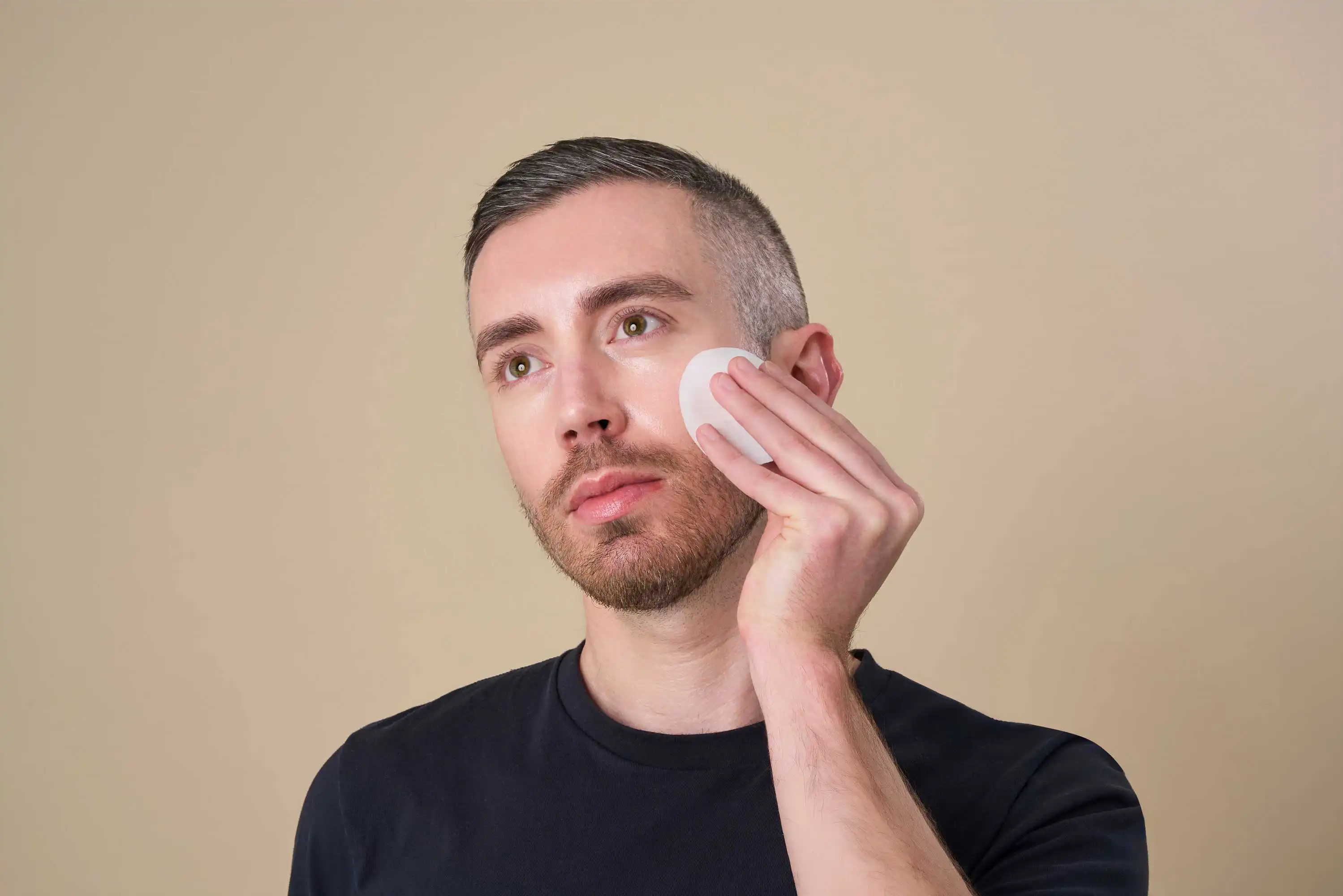 A portrait-style photograph by a Sussex Lifestyle Photographer captures a person with short, dark hair and a trimmed beard gently pressing a white cotton pad against their cheek. Their gaze is directed slightly upwards and to the left. The person is wearing a dark navy or black t-shirt. The background is a plain, light beige or neutral tone, keeping the focus entirely on the individual and the action of skincare application. The lighting is soft and even, highlighting the texture of the skin and the cotton pad.