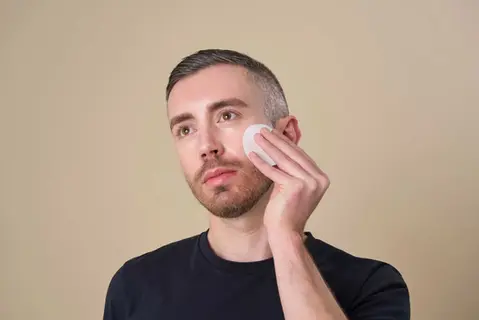 A stylish male model in a black shirt poses confidently against a neutral backdrop, applying an acid wipe to his skin. Captured by a Brighton model photographer in Sussex. This image highlights modern portrait photography and professional branding expertise.