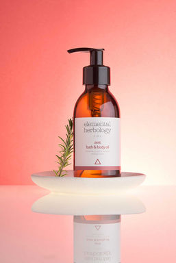 A product shot by a product photographer in Sussex - Brighton showcases Elemental Herbology Zest Bath & Body Oil. Photographed with a still life aesthetic, the amber glass bottle with a black pump dispenser sits on a luminous, stacked white circular platform against a gradient backdrop transitioning from soft peach to a muted coral hue. A sprig of fresh rosemary rests gently beside the bottle, adding a natural, aromatic touch. The white label on the bottle clearly displays "elemental herbology" in elegant lettering at the top, followed by "zest" in a smaller, italicized font. Below, "bath & body oil" is printed, and a minimalist triangle logo in a dusty rose color completes the design. The glossy surface beneath the platform reflects the scene, enhancing the sense of depth and highlighting the product's smooth packaging. This image captures the essence of natural skincare, likely envisioned by a skilled Brighton product photographer like Stuart Price, emphasizing the product's purity and sensory appeal.