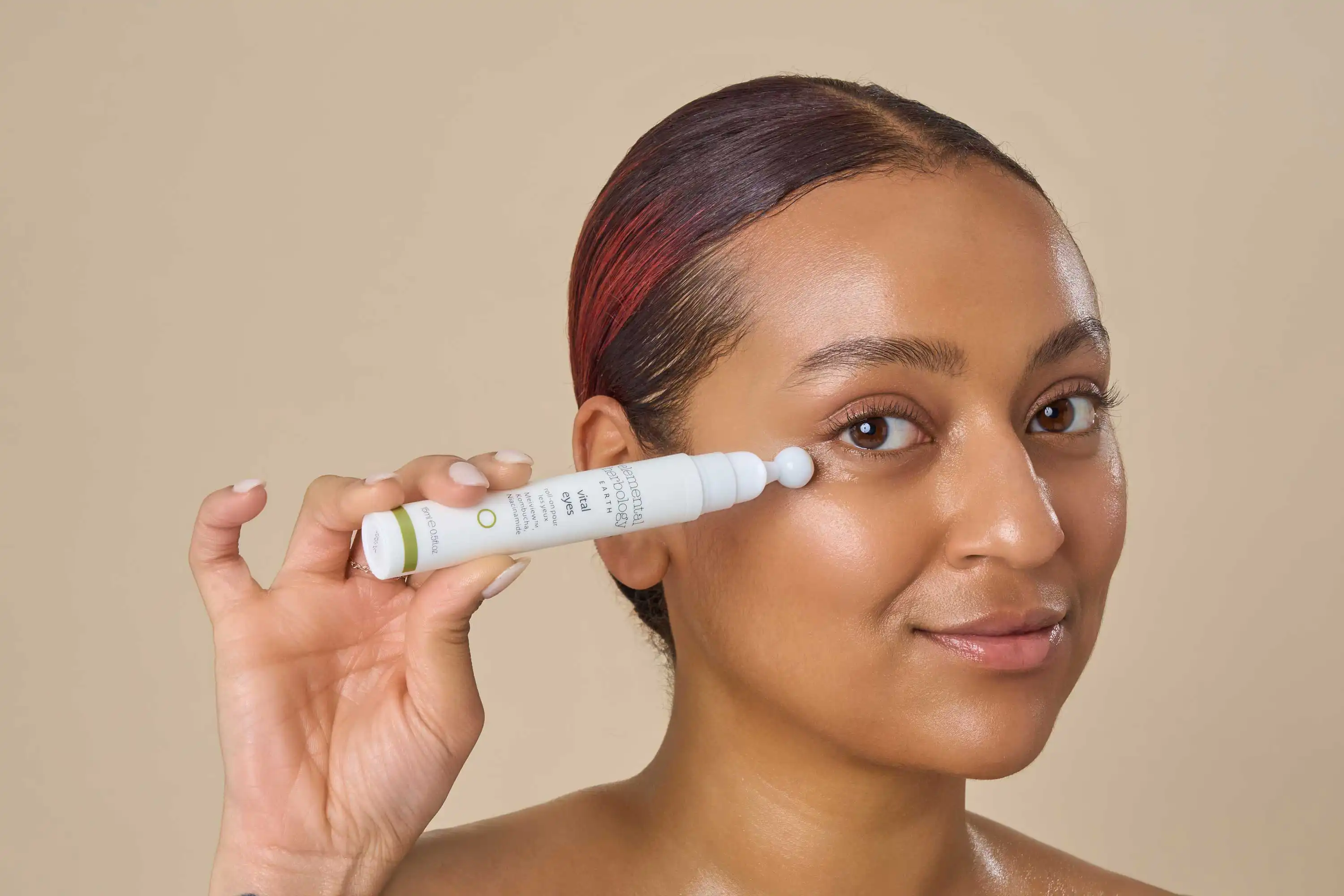 A close-up portrait by a Sussex Lifestyle Photographer captures a person gently applying a skincare product under their eye using a white tube with a rollerball applicator. The person is holding the tube in their left hand, bringing it close to their face. Their gaze is soft and slightly directed towards the camera. The background is a plain, warm beige tone, ensuring the focus remains on the person and the skincare application. The lighting is soft and even, highlighting the texture of the skin and the product.
