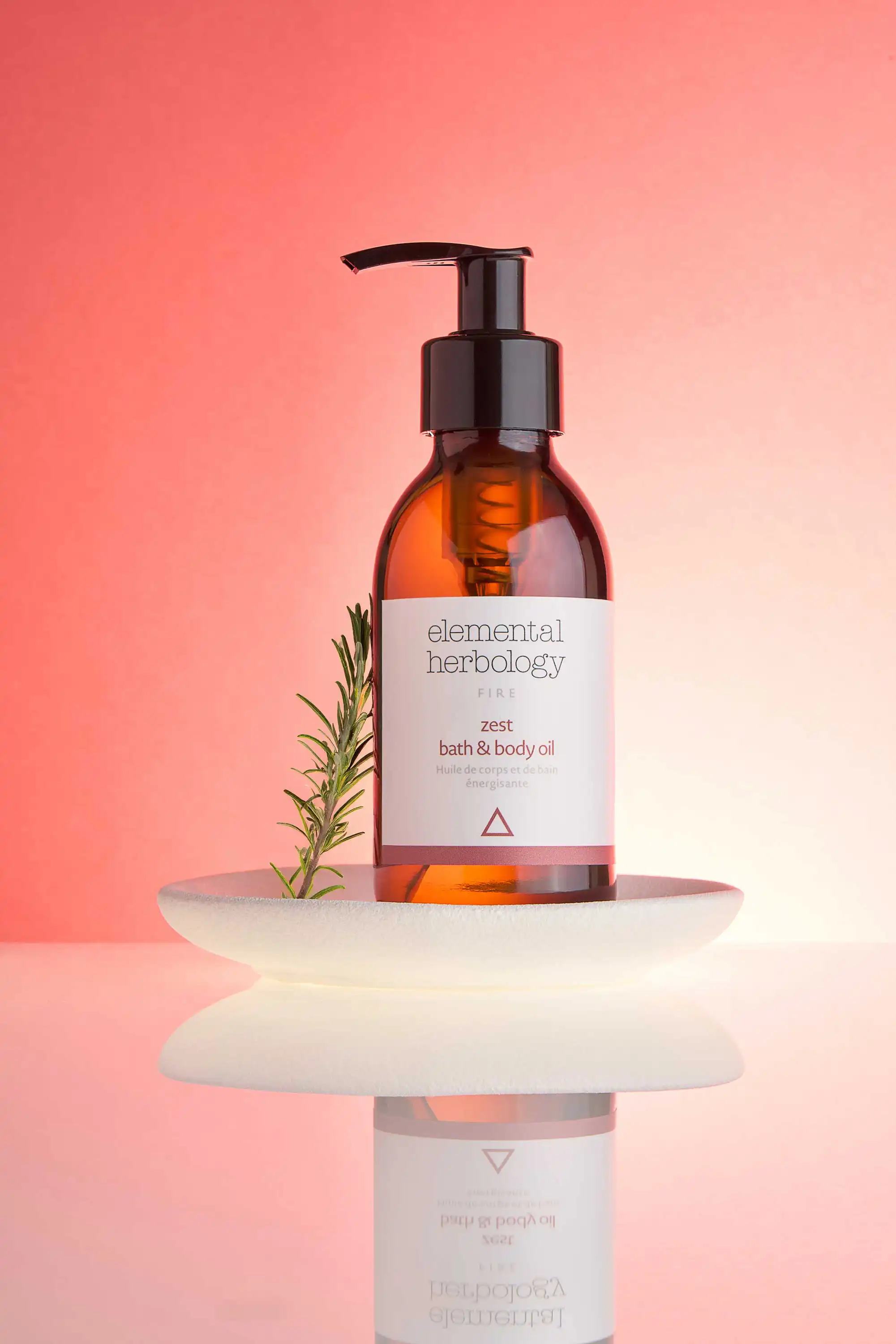 A product photograph by a Sussex product Photographer showcases a clear, amber glass bottle of Elemental Herbology Zest Bath & Body Oil. The bottle has a black pump dispenser and a white label with black and red text. It rests on a stack of two smooth, white, asymmetrical dishes, which create a subtle reflection on the glossy white surface below. A sprig of fresh rosemary leans against the side of the bottle. The background is a gradient of warm pink and peach tones, creating a soft and inviting atmosphere. The lighting is carefully controlled to highlight the product's texture and the minimalist elegance of the presentation.