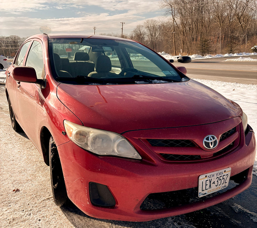 Red Toyota car parked on snowy road, New York license plate visible. Clear winter day with bare trees in the background.