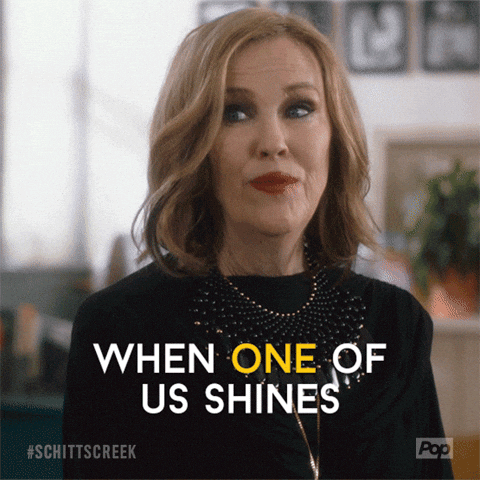 a moving picture of a woman with shoulder length blonde hair in a black dress with a large statement necklace titlting her head saying "when one of us shines all of us shine."