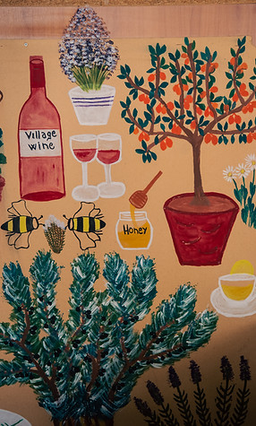 "Colorful hand-painted mural depicting village wine, honey, bees, herbs, a fruit tree, and a cup of tea—celebrating tradition