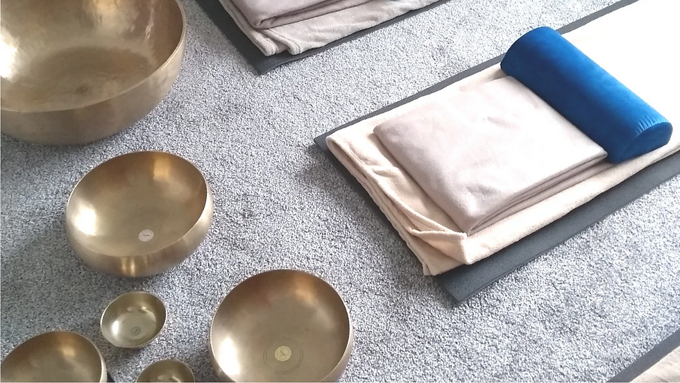 Several singing bowls of different sizes arranged on a grey carpet next to a yoga mat, folded blanket and blue bolster, set up for a Vibration Massage session