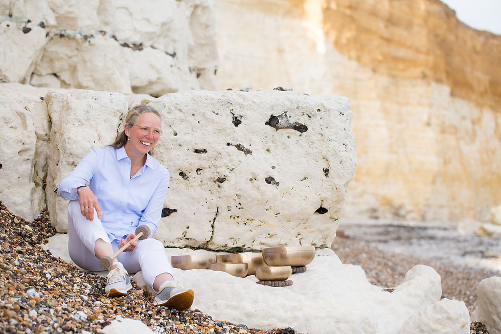 Maja Dethloff sitting on the pebble beach at Seaford, leaning against chalk cliff rocks, with three Peter Hess® singing bowls beside her.