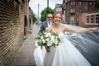 Wind blowing brides veil whilst groom walks behind hetr
