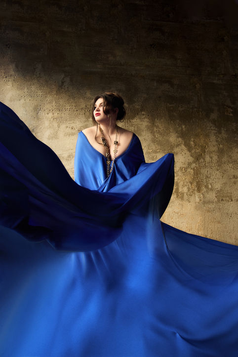 Kseniia Nikolaieva in a flowing royal blue gown, dramatic artistic portrait against a rustic wall.