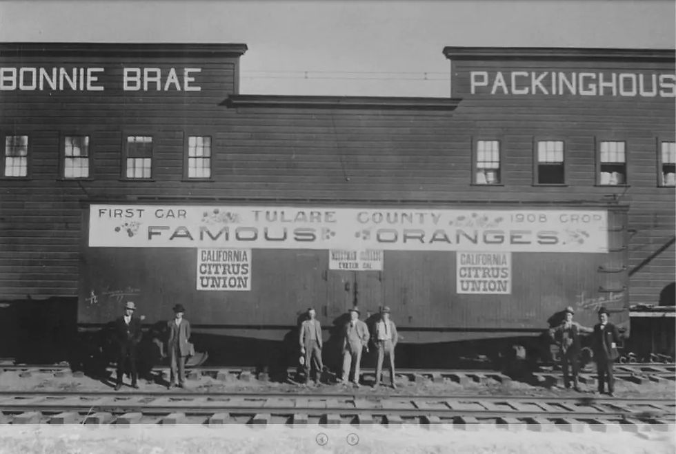 Men stand in front of a train car advertising "Tulare County Famous Oranges" with "Bonnie Brae Packinghouse" in the background, 1908.