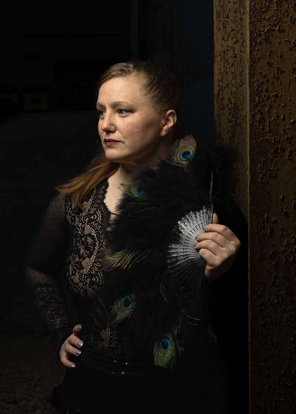 Alt text: Woman in black lace poses with feather fan, gazing off-camera. Rusty pillar and alley in background.